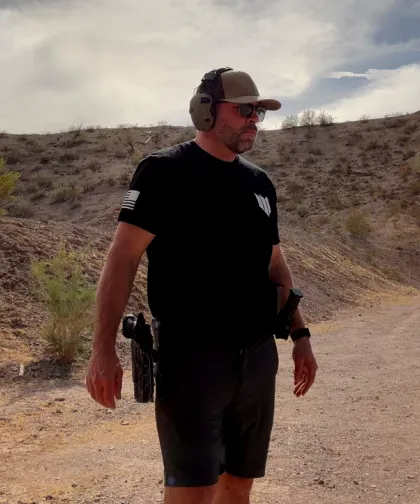 Coach Gregg outdoors at a shooting range, wearing ear protection, a cap and sunglasses